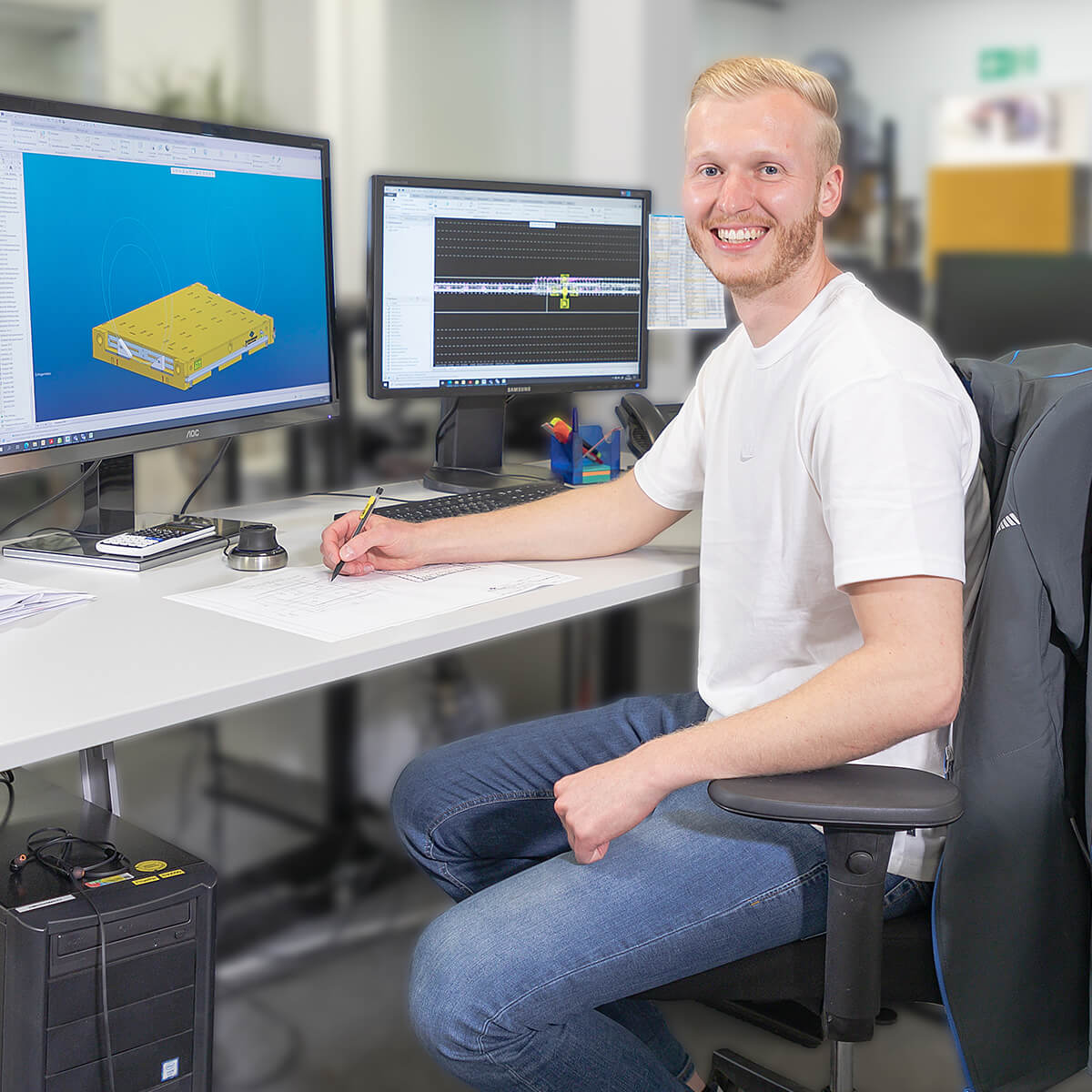 Ein lächelnder Mann sitzt an einem Schreibtisch mit zwei Monitoren. Auf einem Bildschirm ist ein 3D-Modell zu sehen, während der andere Bildschirm Daten anzeigt. Er hält einen Stift und schreibt auf ein Blatt Papier.