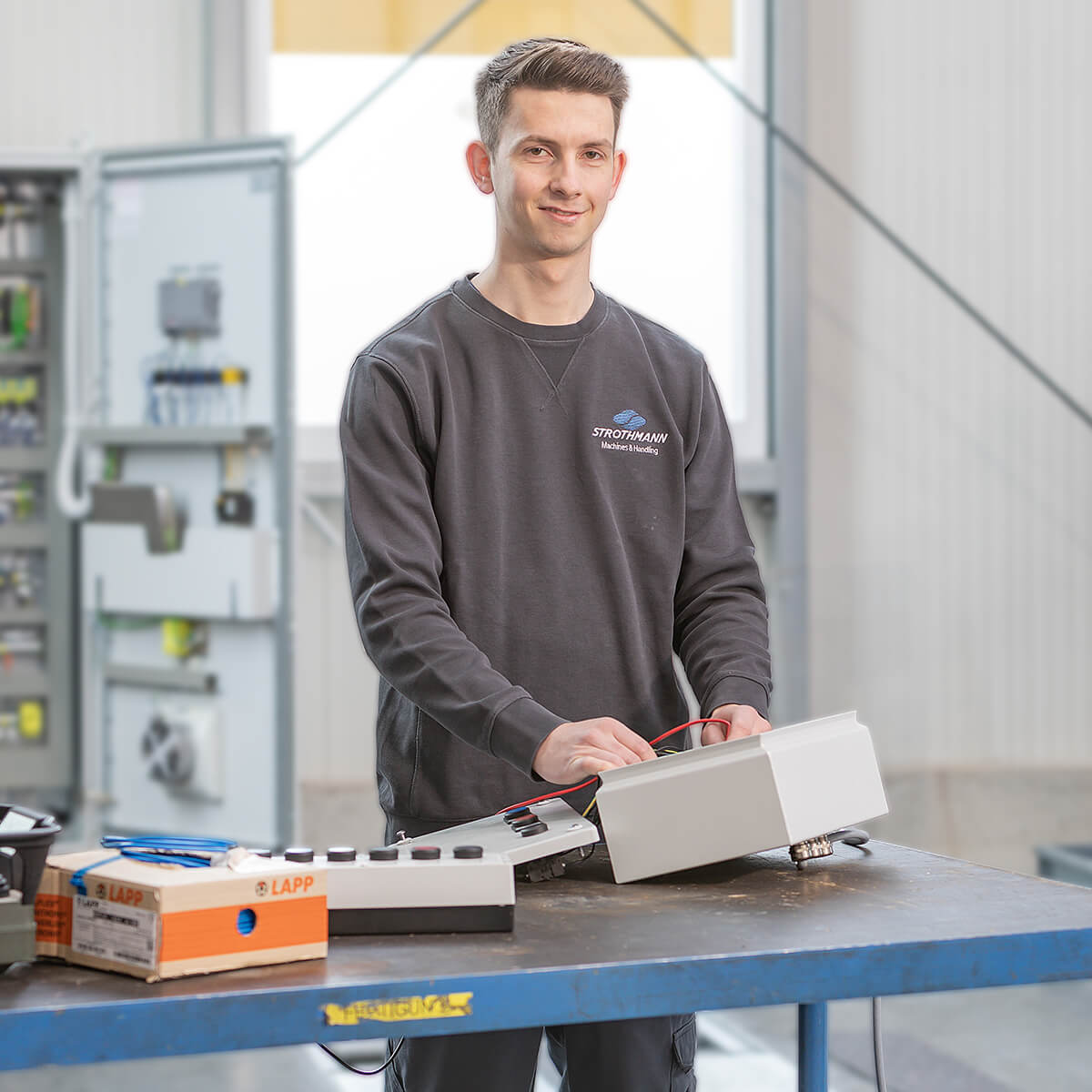 Ein junger Mann in einem Sweatshirt steht an einem Arbeitstisch und arbeitet mit einer elektrischen Steuerung. Im Hintergrund sind Schaltschränke zu sehen. Der Tisch ist mit Kabeln und einer LAPP-Box ausgestattet.