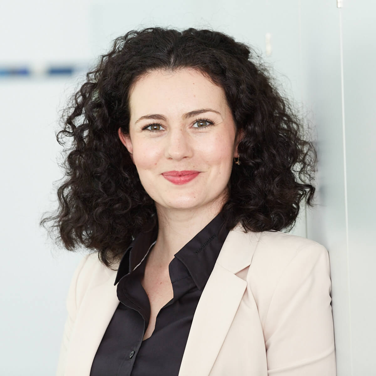 Portrait einer Frau mit lockigem Haar, die in einem hellen Blazer und schwarzem Hemd lächelt. Der Hintergrund ist unscharf und hell.