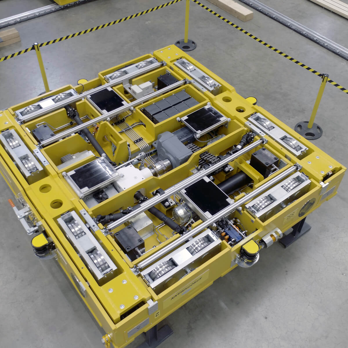 Eine gelbe, quadratische Maschine mit zahlreichen mechanischen Komponenten und Sensoren, in einer Fabrikumgebung.